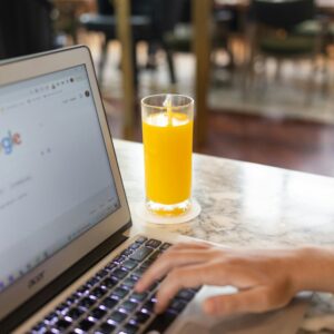 Woman typing on laptop with Google homepage open, beside a glass of orange juice on a marble table.