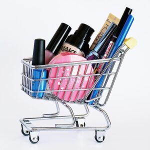 Miniature shopping cart filled with a variety of cosmetics on a white background.