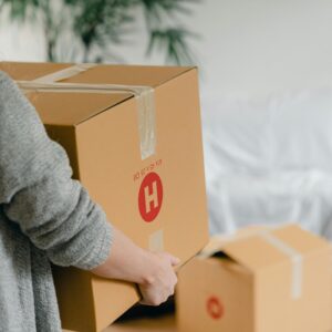 Side view of a woman carrying a cardboard box in a cozy living room, symbolizing moving or relocation.